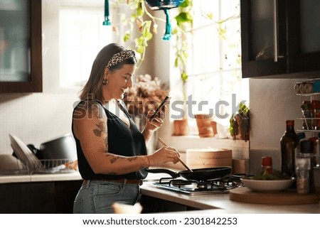 Happy, woman cooking food with smartphone and in kitchen of a home. Dinner or lunch, multitasking and plus size, natural person with tattoo on cellphone following recipe motivation for healthy diet