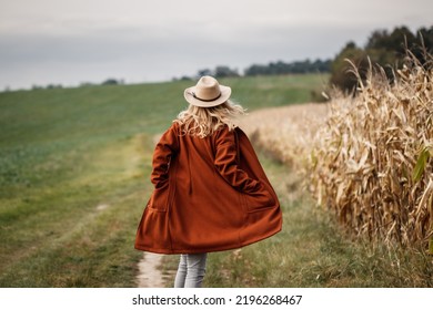 Happy Woman With Cowboy Hat And Red Coat Running At Corn Field. Fall Season In Countryside. Autumn Fashion Collection