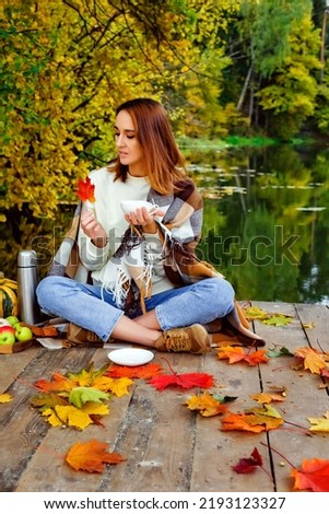 Happy woman in cozy knitted sweater drinking tea from thermos. She is sitting near lake in autumn forest.