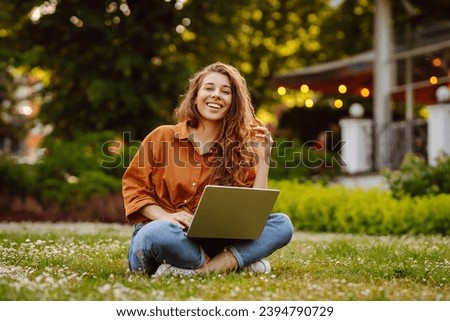 Happy woman with curly hair with a laptop on a green meadow at sunset. Young freelancer woman working outdoors. Freelance work, nature concept.
