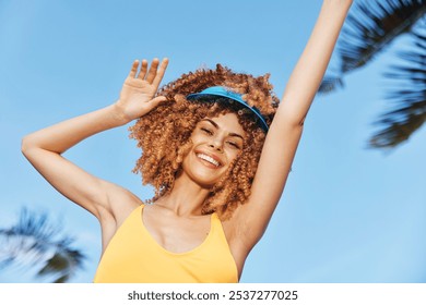 Happy woman with curly hair wearing a yellow swimsuit and visor, enjoying a sunny day outdoors against a clear blue sky - Powered by Shutterstock