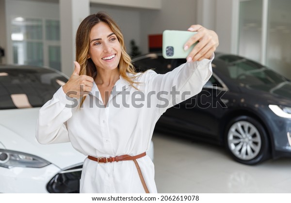 Happy woman customer buyer client in white shirt\
do selfie shot mobile cell phone show thumb up choose auto want to\
buy new automobile in car showroom vehicle salon dealership store\
motor show indoor.