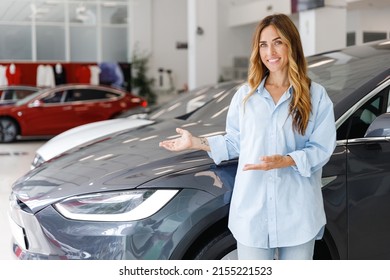 Happy Woman Customer Female Buyer Client In Blue Shirt Point Hands Arms On Dark Car Choose Auto Want To Buy New Automobile In Showroom Vehicle Salon Dealership Store Motor Show Indoor. Sales Concept