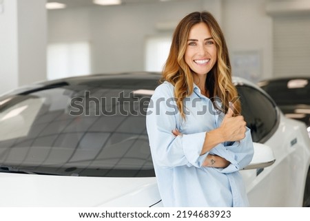 Happy woman customer female buyer client in blue shirt leaning on hood show thumb up choose auto want buy new automobile in car showroom vehicle salon dealership store motor show indoor. Sales concept