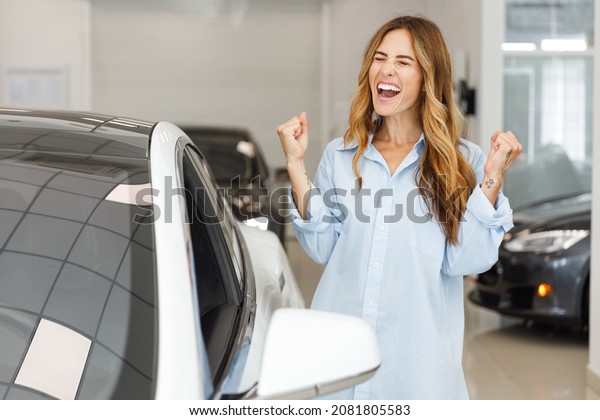 Happy woman customer female buyer client wears\
blue shirt do winner gesture chooses auto wants to buy new\
automobile in car showroom vehicle salon dealership store motor\
show indoor. Sales\
concept.