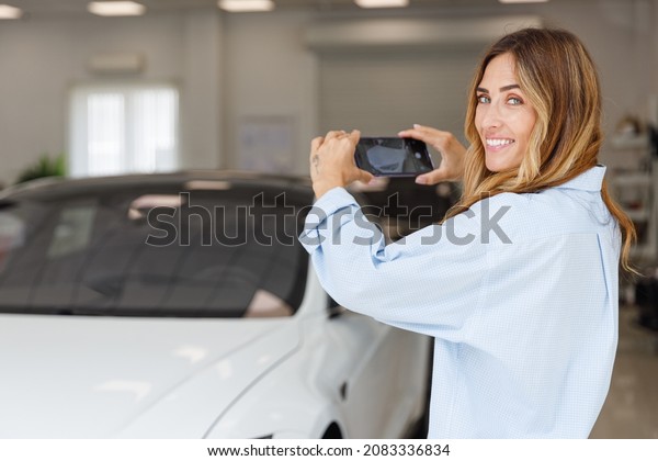Happy woman customer female buyer client wears\
blue shirt do photo on mobile phone choose auto want buy new\
automobile in car showroom vehicle salon dealership store motor\
show indoor. Sales\
concept