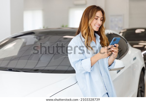 Happy woman customer female buyer client wears\
blue shirt use mobile cell phone choose auto want to buy new\
automobile in car showroom vehicle salon dealership store motor\
show indoor. Sales\
concept