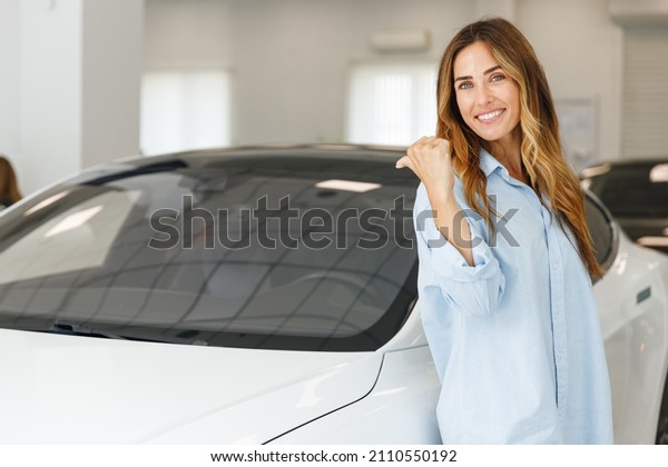 Happy woman customer female buyer client wears\
blue shirt chooses point finger on white auto wants to buy new\
automobile in car showroom vehicle salon dealership store motor\
show indoor. Sales\
concept