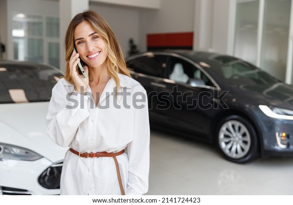 Happy woman customer female buyer client in white\
shirt talk on mobile cell phone choose auto want to buy new\
automobile in car showroom vehicle salon dealership store motor\
show indoor. Sales\
concept
