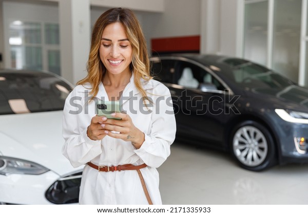Happy woman customer female buyer client in white\
shirt using mobile cell phone choose auto want to buy new\
automobile in car showroom vehicle salon dealership store motor\
show indoor. Sales\
concept