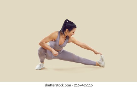 Happy Woman Doing Leg Stretching Exercise During Her Fitness Workout At Gym. Beautiful Flexible Athletic Lady In Yoga Pants Isolated On Beige Color Background Reaching Toward One Foot With Her Hand