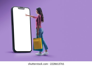 Happy woman doing online shopping using a big smartphone, she is holding shopping bags and touching the blank screen
