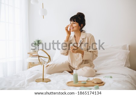 Happy woman doing routine skin care at home with beauty products. Woman sitting on bed at home and applying face cream.