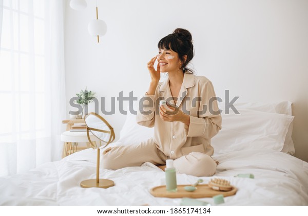 Happy\
woman doing routine skin care at home with beauty products. Woman\
sitting on bed at home and applying face\
cream.