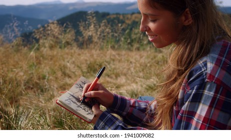 Happy woman drawing sketch of trees in notebook at weekend in mountains. Positive girl painting picture with pen. Smiling female hiker enjoying natural landscape. Young lady taking rest during hike