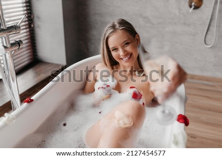 Happy woman drinking champagne while lying in foamy bathtub, celebrating holiday with alcoholic beverage and stretching glass to camera. Young lady relaxing in hot water at luxury hotel