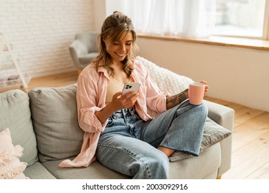 Fröhliche Frau trinkt am frühen Morgen Kaffee, sitzend auf dem Sofa in rosafarbenem Hemd zu Hause. Mit Smartphone. 