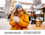 Happy woman drinks a hot drink at a Christmas market decorated with festive lights. Festive Christmas fair, winter holidays concept.