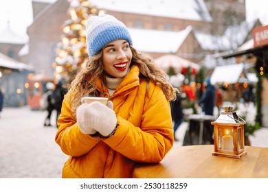 Happy woman drinks a hot drink at a Christmas market decorated with festive lights. Festive Christmas fair, winter holidays concept. - Powered by Shutterstock