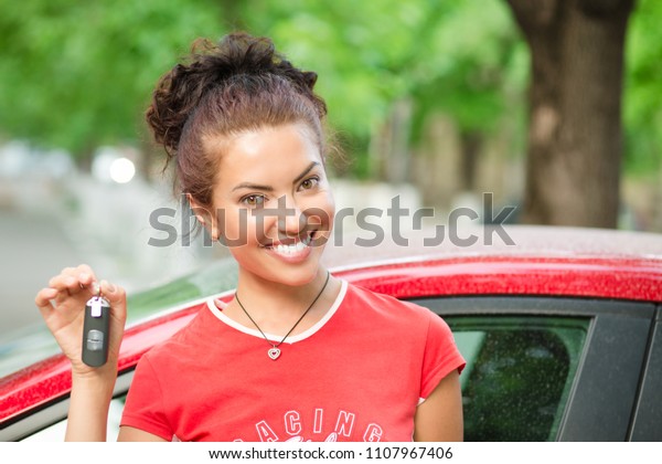 Happy woman driver showing car keys and leaning on\
car door wearing red shirt outdoors, outside in summer day in city\
building and green trees on background. Mixed race african american\
caucasian braz