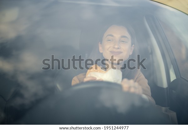 Happy woman eating a burger in the car while\
driving and looking at road. Have unhealthy fast food snack. Food\
to go. Hungry and busy\
concept.