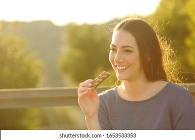 Happy Woman Eating A Cereal Bar Sitting On A Bench At Sunset In A Park