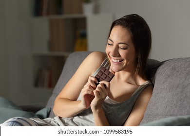 Happy Woman Eating Chocolate In The Night Sitting On A Couch In The Living Room At Home