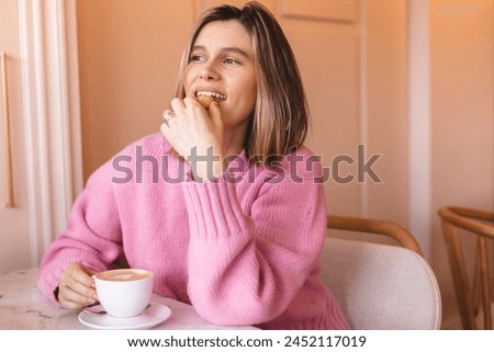 Happy woman eating cookie sitting in a restaurant. Positive woman bites cookie enjoys eating sweet food breaks diet looks away. Woman wear pink sweater rest in cafe. Girl bite cake and look happy.