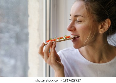 Happy Woman Eating Rye Crisp Bread With Creamy Vegetarian Cheese Tofu, Cherry Tomato And Rucola Micro Greens, Sitting At Home And Looking At Window. Healthy Food, Gluten Free, Diet Concept. 