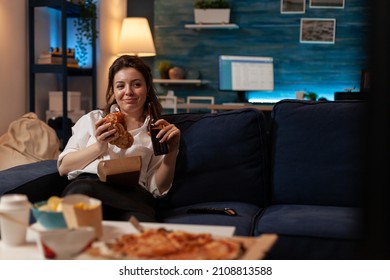 Happy Woman Eating Tasty Takeaway Hamburger Holding Beer Bottle Relaxing On Couch Watching Television Family Sitcom. Smiling Person Having Takeout Burger And Fast Food Delivery Menu.