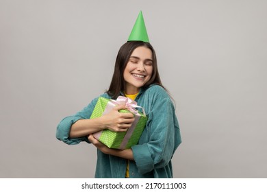 Happy Woman Embracing Gift Box And Smiling With Pleasure Expression, Hugging With Love Enormous Birthday Present, Wearing Casual Style Jacket. Indoor Studio Shot Isolated On Gray Background.