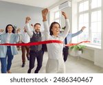 Happy woman employee running to the red ribbon with raised arms celebrating crossing finishing line in office with people colleagues in background. Business competition and teamwork concept.