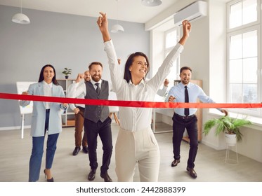 Happy woman employee running to the red ribbon with raised arms celebrating crossing finishing line in office with people colleagues in background. Business competition and teamwork concept. - Powered by Shutterstock