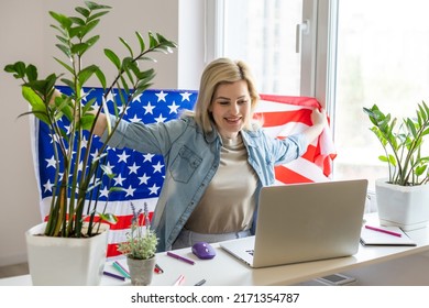 Happy woman employee sitting wrapped in USA flag, shouting for joy in office workplace, celebrating labor day or US Independence day. Indoor studio studio shot isolated on yellow background