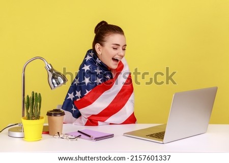 Happy woman employee sitting wrapped in USA flag, shouting for joy in office workplace, celebrating labor day or US Independence day. Indoor studio studio shot isolated on yellow background.