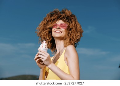 Happy woman enjoying ice cream on a sunny day, wearing pink sunglasses and a yellow swimsuit, showcasing summer vibes - Powered by Shutterstock