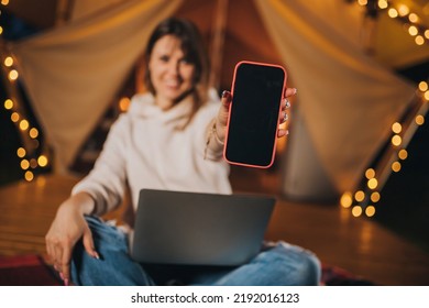 Happy Woman Freelancer Showing Phone While Working Laptop On Cozy Glamping Tent In Summer Night. Luxury Camping Tent For Outdoor Holiday And Vacation. Lifestyle Concept