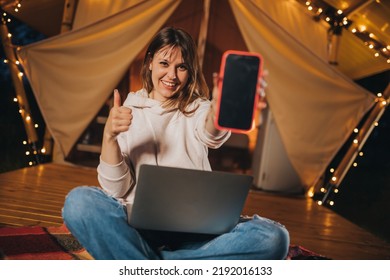 Happy Woman Freelancer Showing Phone While Working Laptop On Cozy Glamping Tent In Summer Night. Luxury Camping Tent For Outdoor Holiday And Vacation. Lifestyle Concept