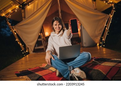 Happy Woman Freelancer Showing Phone While Working Laptop On Cozy Glamping Tent In Summer Night. Luxury Camping Tent For Outdoor Holiday And Vacation. Lifestyle Concept