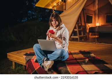 Happy Woman Freelancer Using Phone And Working Laptop On Cozy Glamping Tent In Summer Night. Luxury Camping Tent For Outdoor Holiday And Vacation. Lifestyle Concept