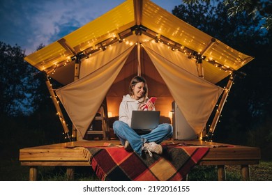 Happy Woman freelancer using phone and working laptop on cozy glamping tent in summer night. Luxury camping tent for outdoor holiday and vacation. Lifestyle concept - Shutterstock ID 2192016155