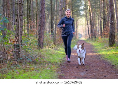 Happy woman full of vitality exercising her dog as they run together along a footpath through forest trees in a healthy active lifestyle concept