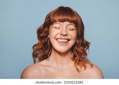 Happy woman with ginger hair and flawless skin smiling with her eyes closed. Cheerful young woman embracing her natural beauty. Body confident young woman standing against a blue studio background. - Powered by Shutterstock