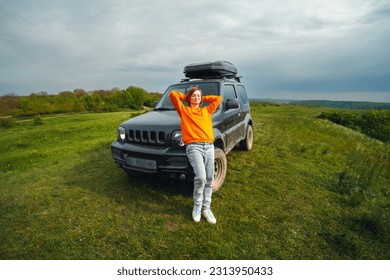 A happy woman, a girl in a bright orange sweater, is standing next to a small SUV with a roof rack. View point on the top of the mountain, Travel and tourism concept.