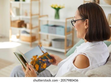 Happy woman in glasses sitting on sofa at home and looking through pictures in printed family album. Smiling lady enjoying good memories while looking through her hardcover photo book. Copy space blur