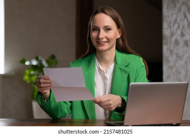 Happy Woman In Green Jacket Reads Acceptance Letter From University Smiling. Young Female Student Sits At Wooden Desk Near Open Laptop At Home