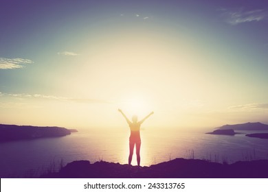 Happy woman with hands up standing on cliff over sea and islands at sunset. Vintage mood, concepts of winner, freedom, happiness etc.