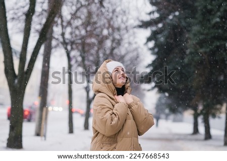 A happy woman in a hat is warming herself in a coat. It stands in the city in winter under the snow