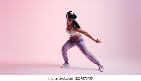 Happy woman having fun in the metaverse. Active young woman gaming with virtual reality goggles and controllers. Energetic young woman enjoying a 3D experience in a studio. - Powered by Shutterstock