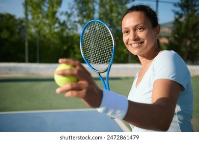 Happy woman in her 40s, enjoying a game of tennis on an outdoor court. She is holding a tennis ball and racket, wearing a white wristband, and smiling with joy and confidence. - Powered by Shutterstock